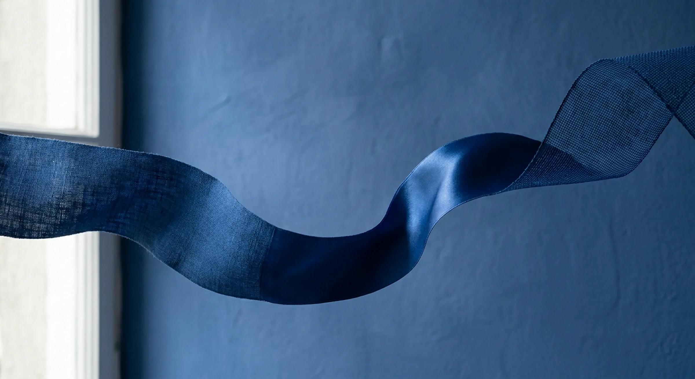 A single ribbon of cobalt fabric sweeping across a plaster wall, its weave changing along its length from raw linen to satin to open mesh.